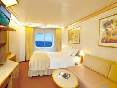 Helle Außenkabine der Carnival Spirit. Doppelbett, beiges Sofa, TV und Fenster mit weitem Meerblick. Warme Holztöne und helle Stoffe im gemütlichen Innenraum.