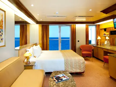 Blick in eine helle und moderne Balkonkabine auf der Carnival Spirit. Zu sehen sind ein gemachtes Doppelbett, ein Sofa, ein Schreibtischbereich mit TV und ein Balkon mit weitem Meerblick.
