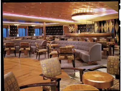 Blick in eine weitläufige Schiffsbar oder Lounge mit mehreren Sitzbereichen. Elegante Sessel und Sofas umgeben goldene Tische. Im Hintergrund eine Bar mit hohen Hockern und einer kunstvollen Wandverkleidung aus Rautenmustern. Die Decke ist ebenfalls in glänzendem Gold gehalten.