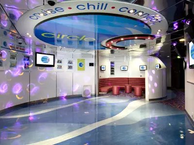 Innenansicht eines modernen Jugendbereichs an Bord der Carnival Spirit. Der Raum hat einen blauen und weißen Boden, der an Wellen erinnert, rote gepolsterte Sitzgelegenheiten, Bildschirme mit dem 'Circle C'-Logo und eine 'Wall of Fame' für 12-14-Jährige. Violette Lichtreflexe tanzen an den Wänden und dem glänzenden Boden.