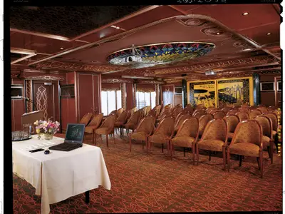 Ein leerer, klassisch eingerichteter Konferenzraum auf der Carnival Spirit mit Reihen von gepolsterten Holzstühlen. Vorne ein Rednerpult mit Mikrofon und Laptop. Die Decke ist mit einer großen, bunten Buntglas-Lichtinstallation und einem Projektor verziert.
