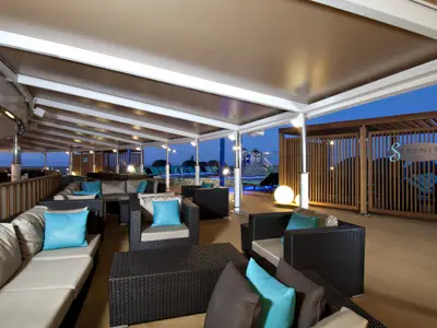 Breite Aufnahme einer überdachten Outdoor-Lounge auf der Carnival Spirit mit gemütlichen Sitzgruppen und Blick auf das Meer in der Dämmerung. Blaue Kissen und warme Beleuchtung sorgen für entspannte Atmosphäre.