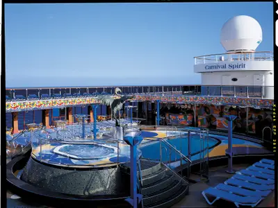 Pooldeck der Carnival Spirit mit bunten Pools, Whirlpool, Liegestühlen und Bar. Eine Kranich-Skulptur ziert die Mitte, dahinter weites blaues Meer.