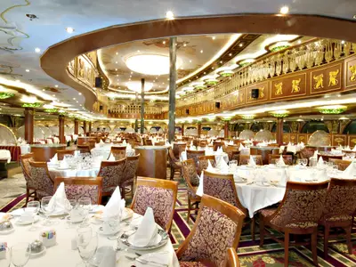 Blick in ein großes, luxuriöses Restaurant auf der Carnival Spirit. Zahlreiche runde Tische sind mit weißen Tischdecken, Gläsern und Besteck festlich gedeckt. Eine obere Galerie mit goldenen Verzierungen umgibt den Raum. Die Decke ist bemalt.