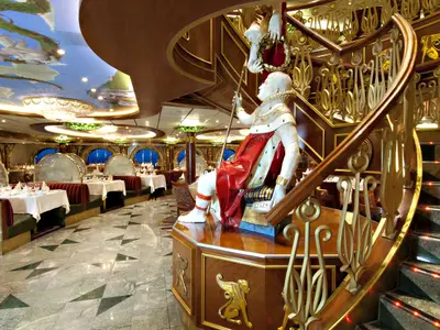 Ein opulent eingerichtetes Schiffsrestaurant mit marmoriertem Boden, elegant gedeckten Tischen und Kronleuchtern. Eine prunkvolle Treppe mit einer Statue im Vordergrund dominiert den Raum. Die Decke ist mit einem gemalten Himmel verziert.
