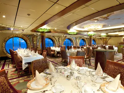 Ein opulentes Restaurant der Carnival Spirit mit runden Bullaugen-Fenstern, die den Blick auf das blaue Meer freigeben. Die Tische sind festlich gedeckt, der Raum ist reich verziert mit goldenen Elementen, gemusterten Teppichen und Kronleuchtern. Keine Personen sind sichtbar.