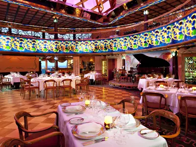 Ein luxuriöses, ovales Restaurant auf der Carnival Spirit. Gedeckte Tische mit weißen Tischdecken und Kerzen sind bereit. Ein Flügel, eine Weinbar und ein Fenster mit Meerblick sind sichtbar. Die Decke ist mit bunten Lichtinstallationen verziert.