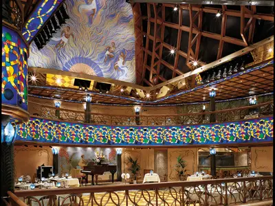 Opulentes Restaurant auf der Carnival Spirit. Die mehrstöckige Halle präsentiert farbenfrohes Buntglas, eine große Wandmalerei mit Figuren und einen Flügel auf einer Bühne. Tische sind eingedeckt.