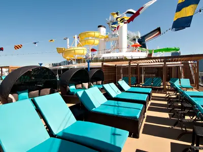 Blick auf das obere Sonnendeck der modernen Carnival Spirit. Mehrere blaue Liegen und geflochtene Cabanas stehen bereit. Im Hintergrund sind gelbe Wasserrutschen und Stadtgebäude zu sehen. Blauer Himmel, tagsüber.