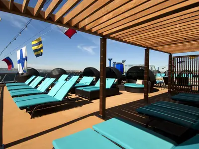 Geräumiges Sonnendeck der Carnival Spirit mit zahlreichen türkisfarbenen Sonnenliegen, schwarzen Cabanas und einer hölzernen Pergola. Im Hintergrund sind Meer und Himmel sichtbar.