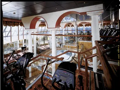 Blick von oben in ein mehrstöckiges Fitness- und Spa-Areal auf der Carnival Spirit mit Trainingsgeräten, Whirlpool, Säulen, Holzböden und großen Fenstern zum Meer.