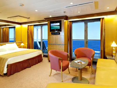 Innenansicht einer geräumigen Suite auf der Carnival Spirit mit Doppelbett, Sitzbereich und Meerblick vom Balkon. Helle, goldene Farbtöne dominieren.