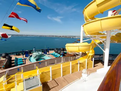 Sonniges Oberdeck der Carnival Spirit mit gelben Wasserrutschen, Pools und Liegestühlen. Blauer Himmel, weites Meer mit Stadtkulisse im Hintergrund. Bunte Flaggen.