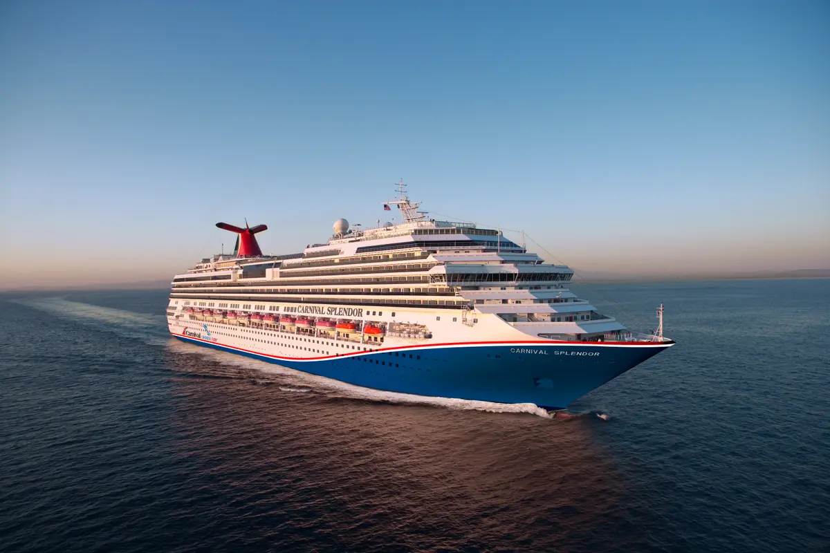 Die große Carnival Splendor mit blauem Rumpf und weißem Aufbau fährt bei Sonnenuntergang auf dem offenen Meer, das klare Wasser schäumt am Bug.