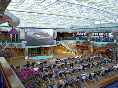 Großer, heller Atrium-Bereich auf der Carnival Splendor unter einem Glasdach. Viele Gäste entspannen auf Liegestühlen vor einer großen Videoleinwand, die ein Schiff zeigt. Mehrere Ebenen mit Bars und Pflanzen. Blick auf das Meer.
