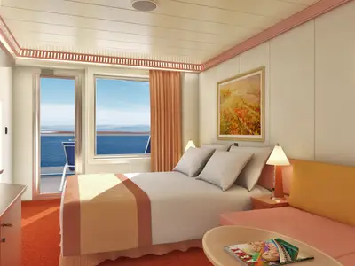 Eine moderne Balkonkabine auf der Carnival Splendor. Ein Doppelbett mit weißen Kissen, eine orangefarbene Couch und ein Beistelltisch sind zu sehen. Links eine Theke mit Kaffeemaschine. Durch die Balkontür blickt man auf das blaue Meer und entfernte Berge.