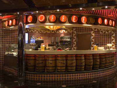 Ein stilvoller Kaffee- und Süßwaren-Shop an Bord der Carnival Splendor mit 'COFFEE SHOP' Neonlicht-Schild über der Theke und dunklen Holzfässern als Tresenverkleidung. Die Wände sind mit gemusterten Fliesen verziert, und es gibt Kaffeemaschinen und Kuchenauslagen zu sehen.