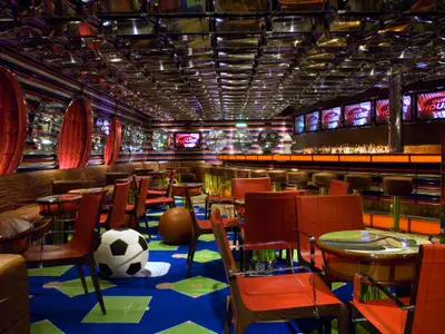 Lebhafte, moderne Bar-Lounge auf der Carnival Splendor mit bunter Einrichtung, Spiegeldecke, Bar, Bildschirmen und Sportbällen.