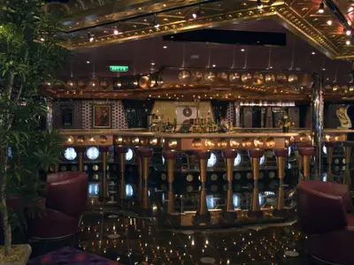 Blick in eine moderne Schiffsbar. Eine lange Theke mit zahlreichen Barhockern, dahinter Flaschen und Spiegel. Rote Sessel und eine Pflanze im Vordergrund. Glänzender Boden spiegelt die Lichter wider.