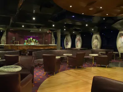 Geräumige, dunkle Lounge auf der Carnival Splendor mit einer gut gefüllten Bar im Hintergrund, vielen bequemen Sesseln und Sofas, kunstvollen Tischen und großen Wandpaneelen mit gemalten Figuren. Der Boden ist teils Parkett, teils Teppich.
