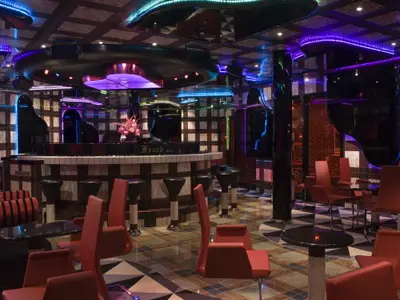 Innenansicht einer stilvollen Bar-Lounge. Eine geschwungene Bar, die einem Klavier nachempfunden ist, steht im Zentrum. Rote Sessel und Barhocker sind vor einer karierten Wand und glänzenden Böden platziert. Blaue und violette LED-Lichter beleuchten den Raum.