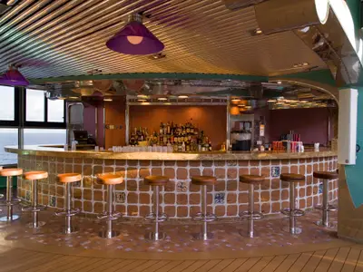 Helle Bar-Lounge auf der Carnival Splendor. Ziegelstein-Bar mit Barhockern, Meerblick durch große Fenster. Viele Spirituosenflaschen, Gläser. Moderne Decke mit violetten Lampen.