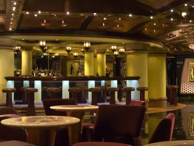 Innenansicht einer eleganten Schiffsbar mit einer geschwungenen Theke, hohen Barhockern und mehreren Säulen. Die Decke ist mit dekorativen Lichtern versehen. Einige Tische und Stühle sind im Vordergrund sichtbar. Die Szene ist warm beleuchtet.