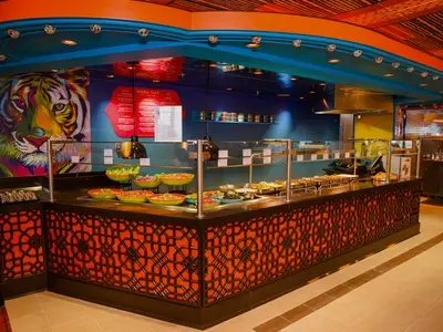Ein lebhaft gestaltetes Buffetrestaurant auf der Carnival Splendor mit einer langen Essensausgabe. Zahlreiche Gerichte in Schalen und Chafing Dishes sind zu sehen. Im Hintergrund dominiert ein farbenfrohes Pop-Art-Gemälde eines Tigers. Decke in Blau und Orange mit dekorativen Lichtern.