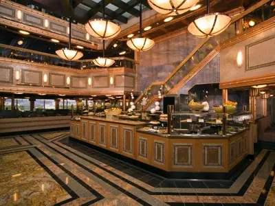 Innenansicht eines eleganten Buffetrestaurants auf der Carnival Splendor. Mehrere Essensstationen mit frischem Obst und Gebäck sind sichtbar. Die Einrichtung ist opulent mit goldenen Akzenten, Mosaikwänden und großen Hängelampen. Im Hintergrund sind Fenster mit Blick nach draußen und weitere Sitzbereiche zu erkennen.