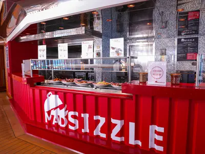 Ein lebhaft roter Mad Sizzle Imbissstand auf der Carnival Splendor. Die Theke präsentiert verschiedene angerichtete Speisen und ein 'Order Here'-Schild. Im Hintergrund sind Speisekarten zu sehen.