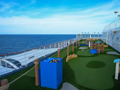 Ein bunter Minigolfplatz an Deck der Carnival Splendor, umgeben von einem Geländer und mit weitem Blick auf das offene Meer und den Horizont unter einem klaren blauen Himmel. Verschiedene Elemente des Minigolfplatzes sind sichtbar, darunter grüne Kunstrasenbahnen und hölzerne Hindernisse.
