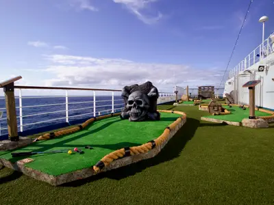 Minigolfplatz im Piraten-Thema auf der Carnival Splendor. Grüne Kunstrasenbahnen mit Hindernissen wie einem großen Totenkopf, einer Schatztruhe und Kanonen. Im Hintergrund ist das blaue Meer und der Himmel zu sehen. Mehrere Golfschläger und Bälle liegen auf der Bahn. Die Begrenzung besteht aus dicken Seilen.