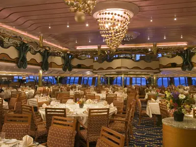 Ein luxuriöser Speisesaal auf der Carnival Splendor, ausgestattet mit zahlreichen gedeckten Tischen, eleganten Stühlen und großen Panoramafenstern, die den Blick auf das offene Meer freigeben. Die Decke und Wände sind aufwendig verziert mit goldenen Elementen und kunstvollen Leuchten. Eine große, goldene Kronleuchter-Skulptur hängt von der Decke.