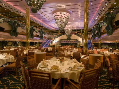 Großzügiges, elegantes Restaurant auf der Carnival Splendor mit vielen runden Tischen, die mit weißen Tischdecken und Geschirr festlich gedeckt sind. Große Kronleuchter und eine geschwungene Treppe sind im Hintergrund sichtbar.