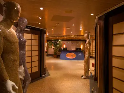 Ein warmer, elegant beleuchteter Flur auf der Carnival Splendor, der zum Spa-Bereich führt. Links und rechts stehen gold- und silberfarbene Mosaik-Mannequins. Im Hintergrund ist ein blauer Empfangstresen mit einem 'Cloud 9 Spa'-Logo zu sehen, flankiert von Pflanzen und warmen Lichtquellen.