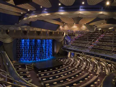 Panoramablick auf ein modernes Theater der Carnival Splendor. Eine blau beleuchtete Bühne mit Vorhang dominiert links, umgeben von geschwungenen Sitzreihen mit kleinen Tischen und weiteren Sitzbereichen auf den Rängen. Elegante Decken- und Wanddekorationen runden das Bild ab.