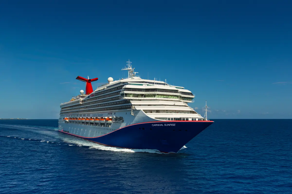 Die Carnival Sunrise fährt auf dem offenen Meer. Blauer Rumpf, weiße Aufbauten und roter Schornstein sind zu sehen. Eine Gischtspur im tiefblauen Wasser unter klarem Himmel ist sichtbar.