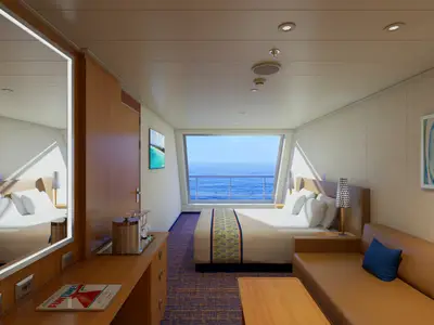Eine helle, moderne Balkonkabine auf der Carnival Sunrise mit Doppelbett, beigefarbenem Sofa, Holztischen und großem Fenster mit Blick auf das weite blaue Meer.