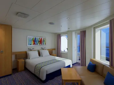 Helle, moderne Balkonkabine mit Doppelbett, Sitzbereich und Holzeinrichtung. Ein farbenfrohes Bild hängt über dem Bett. Durch die Fenster und die Balkontür ist das blaue Meer sichtbar.