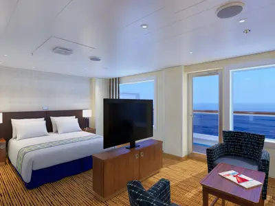 Eine moderne Suite an Bord der Carnival Sunrise. Zu sehen sind ein großes Doppelbett, ein Flachbildfernseher auf einem Holzschrank, ein Sessel und eine Glastür, die zu einem Balkon mit Meerblick führt. Der Ozean und der blaue Himmel sind durch die Fenster sichtbar.