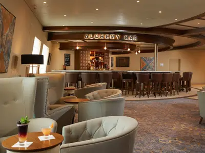Innenansicht der "Alchemy Bar" auf der Carnival Sunrise. Eine lange Theke mit Hockern und bequeme Sessel im Vordergrund. Die Bar ist mit dunklem Holz und Lichtern dekoriert, Kunstwerke an den Wänden.