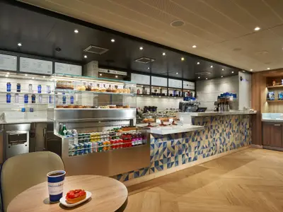 Innenansicht eines modernen Cafés mit hellbraunem Holzboden und einem Tresen, der mit blau-weißen Dreiecksfliesen verkleidet ist. Die Vitrinen sind gefüllt mit Kuchen, Gebäck und verschiedenen Getränken. Im Vordergrund steht ein Tisch mit Kaffee und einem Donut.