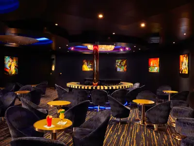 Eine schicke Bar-Lounge auf der Carnival Sunrise bei gedämpftem Licht. Dunkle Samtsessel um goldene Tische. Die Wände sind mit farbintensiven Pop-Art-Bildern dekoriert. In der Mitte befindet sich eine runde Theke mit einem goldenen Pol und einer Piano-Tastatur-Verzierung. Bunte Cocktails stehen auf einem Tisch im Vordergrund.