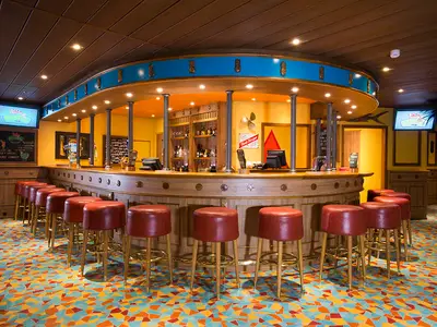 Eine einladende Bar-Lounge auf der Carnival Sunrise mit einer großen ovalen Holztheke, vielen roten Barhockern und einem auffällig bunten Teppich. Im Hintergrund sind Regale mit Flaschen und Bildschirme zu sehen.