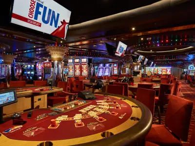 Blick in ein modernes Casino der Carnival Sunrise. Reihen von leuchtenden Spielautomaten und mehrere rote Blackjack-Tische sind sichtbar. Die Decke ist mit bunten Lichtern und Spiegeln verziert. Eine goldene Säule und rote Teppiche prägen die Szene.