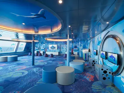 Ein lebhaft gestalteter Kinderbereich auf der Carnival Sunrise mit einem Unterwasser-Thema. Blaue Wände, Decke und Teppich zeigen Meeresmotive. Verschiedene Sitzgelegenheiten und interaktive Bildschirme sind zu sehen. Links befindet sich ein großes Fenster mit Blick auf das Meer.