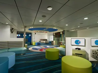 Lebhafter Kinder- und Jugendbereich auf der Carnival Sunrise mit bunten Sitzmöbeln, Kickertisch und Videospielkonsolen. Der Teppichboden zeigt ein Wellenmuster.