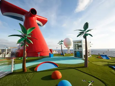 Ein farbenfroher Minigolfplatz auf dem Oberdeck der Carnival Sunrise mit künstlichen Palmen, roten Hindernissen und Blick auf das weite Meer bei Tageslicht.