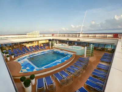 Weitwinkelansicht des Sonnendecks der Carnival Sunrise von einer höheren Ebene. Es zeigt einen Pool mit blauen Kacheln, Liegestühle, einen Whirlpool und einen Bar-Bereich. Das Meer ist im Hintergrund sichtbar.