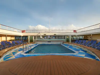 Eine weitwinkel Aufnahme des leeren Hauptpools an Deck der Carnival Sunrise. Blaue Liegestühle säumen das Becken mit blau-weißen Fliesen. Sonniges Wetter, einige Wolken am Himmel. Eine Hebevorrichtung am Beckenrand ist sichtbar.